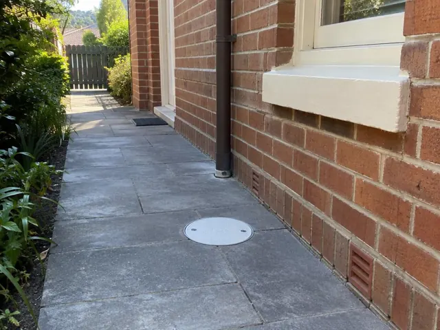 Dry paved side passage beside a brick house with a round drain grate back in place in Cammeray